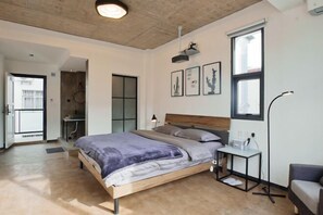 A modern bedroom with a wooden bed, a desk, and a chair. There are framed pictures on the wall and a window with blinds.