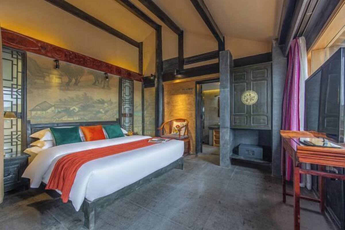 A hotel room with a bed, a chair, a table, and a wall with a mural.