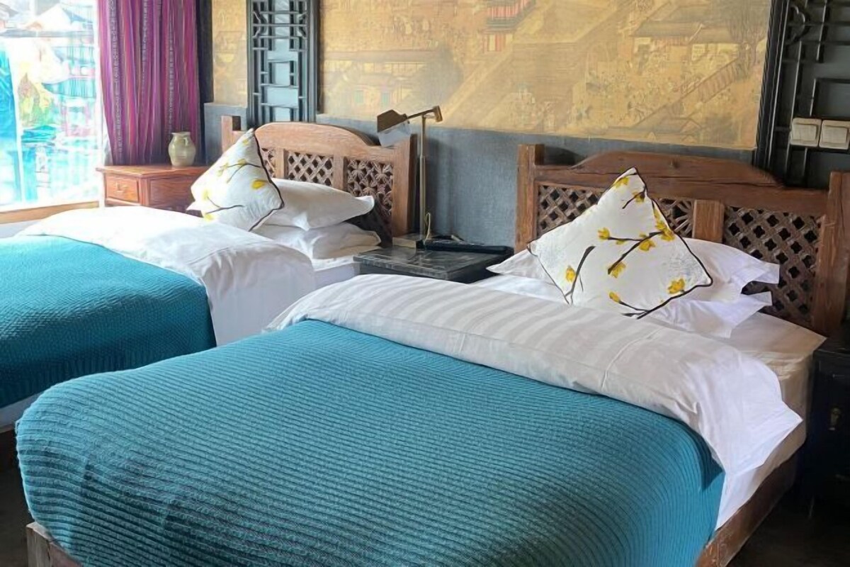 A hotel room with two beds, each with a blue bedspread and white pillows, a wooden headboard, a nightstand, and a wall with a traditional design.