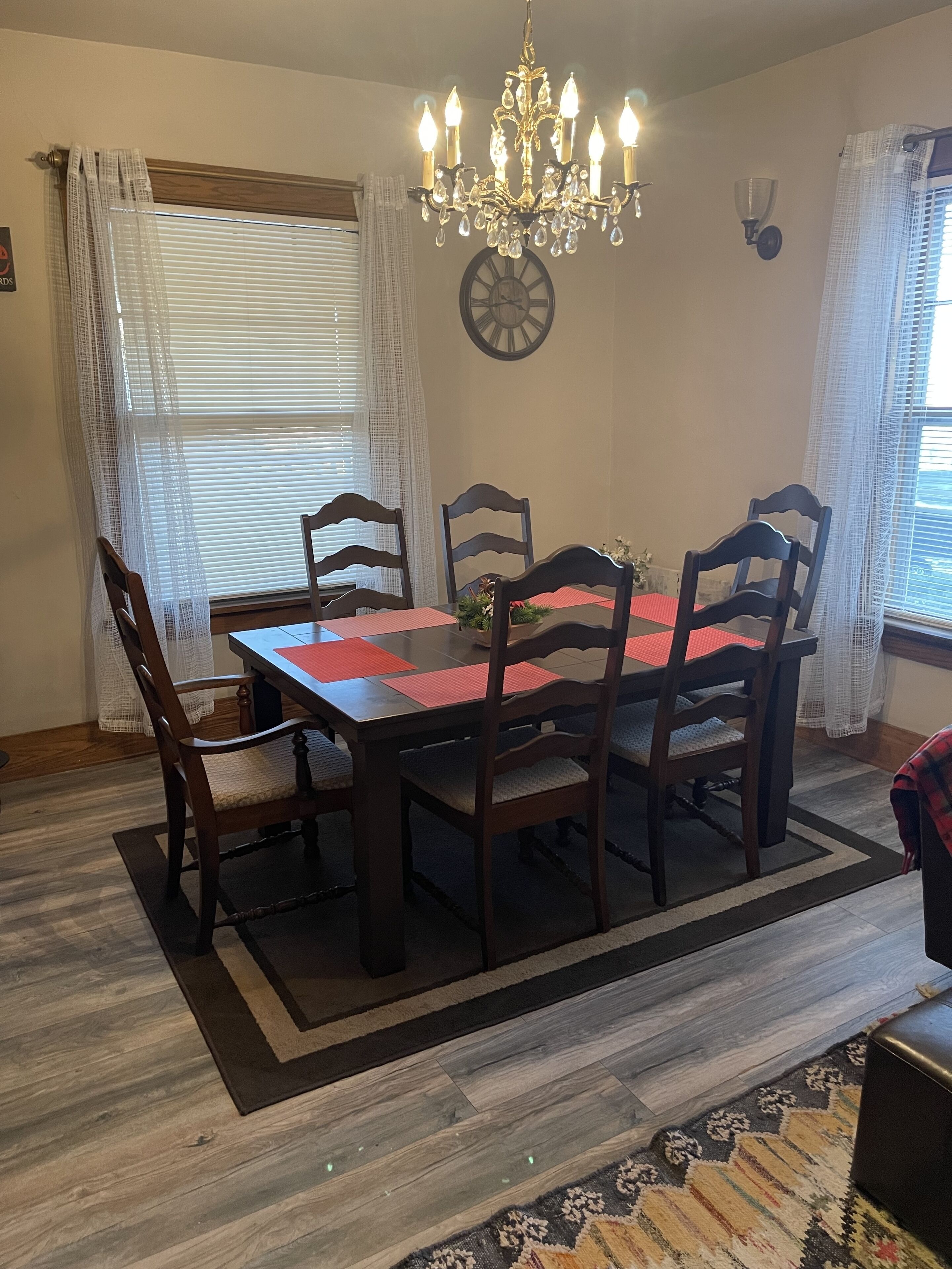 A dining room with a wooden table, six chairs, a chandelier, and a clock on the wall.