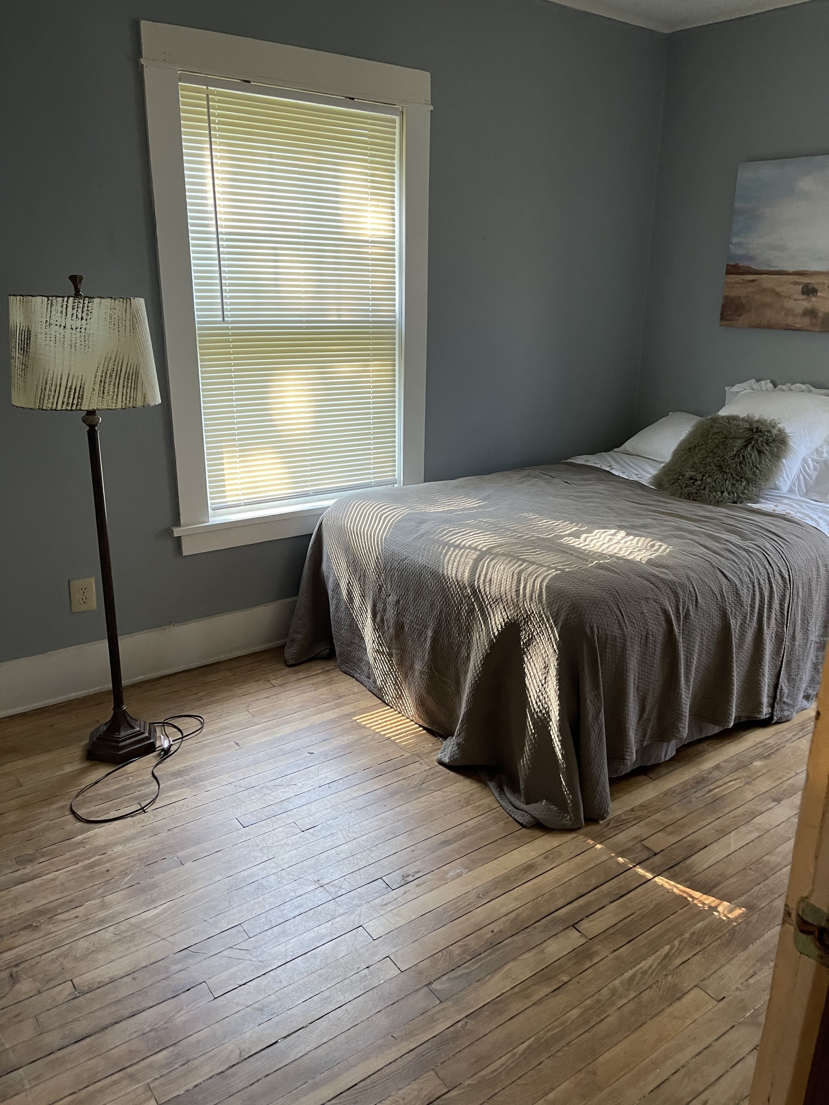 A bedroom with a bed, a floor lamp, a window with blinds, and a painting on the wall.