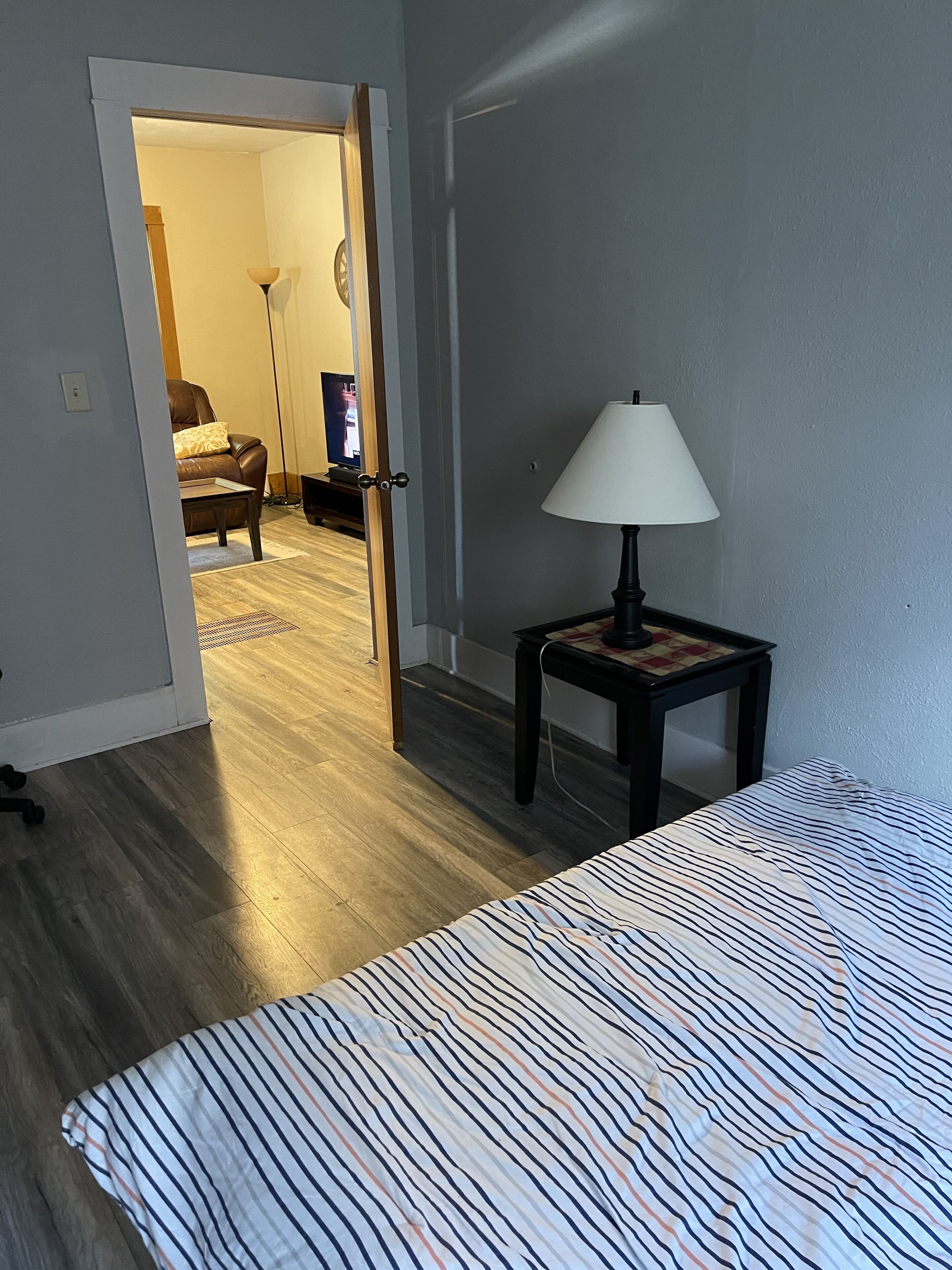 A bedroom with a bed, a nightstand with a lamp, a wooden floor, and a door leading to another room.