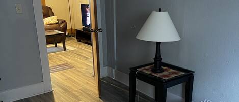 A bedroom with a bed, a nightstand with a lamp, a wooden floor, and a door leading to another room.