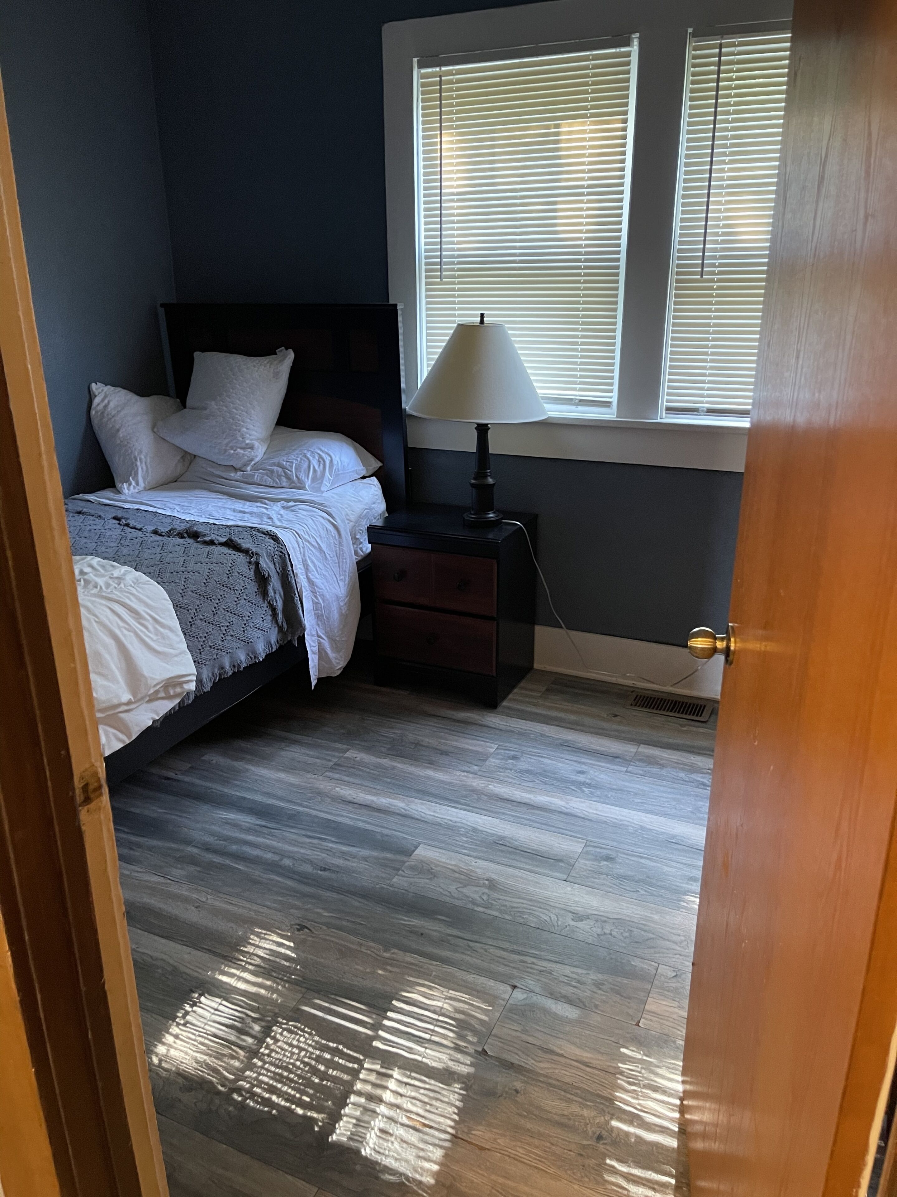 Een slaapkamer met een bed, nachtkastje, lamp en jaloezieën.