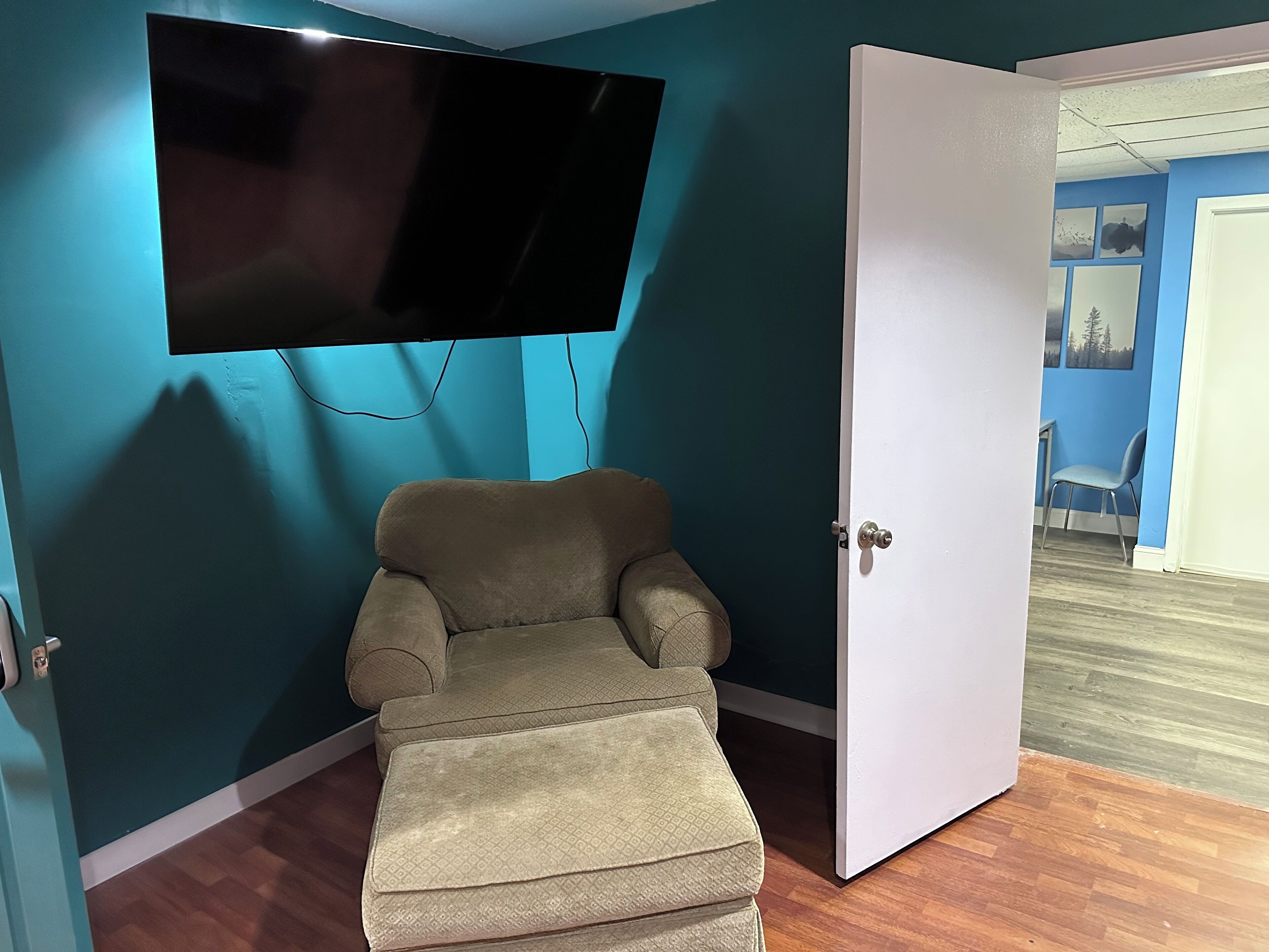 Ein Zimmer mit türkisfarbenen Wänden, einem beigen Sessel, einem an der Wand montierten Flachbildfernseher und einer weißen Tür.