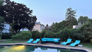Ein Schwimmbecken mit Liegestühlen und einer Sitzecke, umgeben von Grünflächen und Wohnhäusern.