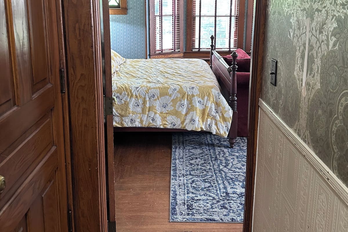 Un couloir étroit mène à une chambre à coucher qui contient un lit, une commode et une fenêtre avec des volets.