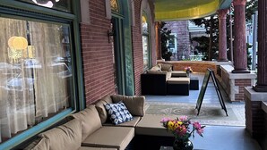 Ein Sitzbereich im Freien mit gepolsterten Bänken und einem Couchtisch, ein großes Fenster mit Vorhängen sowie eine dekorative Vase mit Blumen.