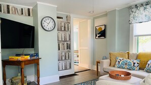Un soggiorno con televisore a schermo piatto, una libreria, un orologio, un divano con cuscini decorati e una finestra con tende.