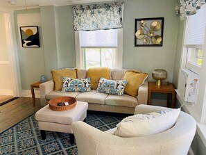 Un salon comprenant un canapé, un pouf et un fauteuil, un tapis à motifs, une fenêtre avec des rideaux et une lampe fixée au mur.