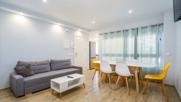 A modern living room with a grey sofa, white coffee table, and a dining area with a white table and yellow chairs. - WELCS APARTAMENTO 027 EMP en el puerto de Roses (Rosas / Roses)