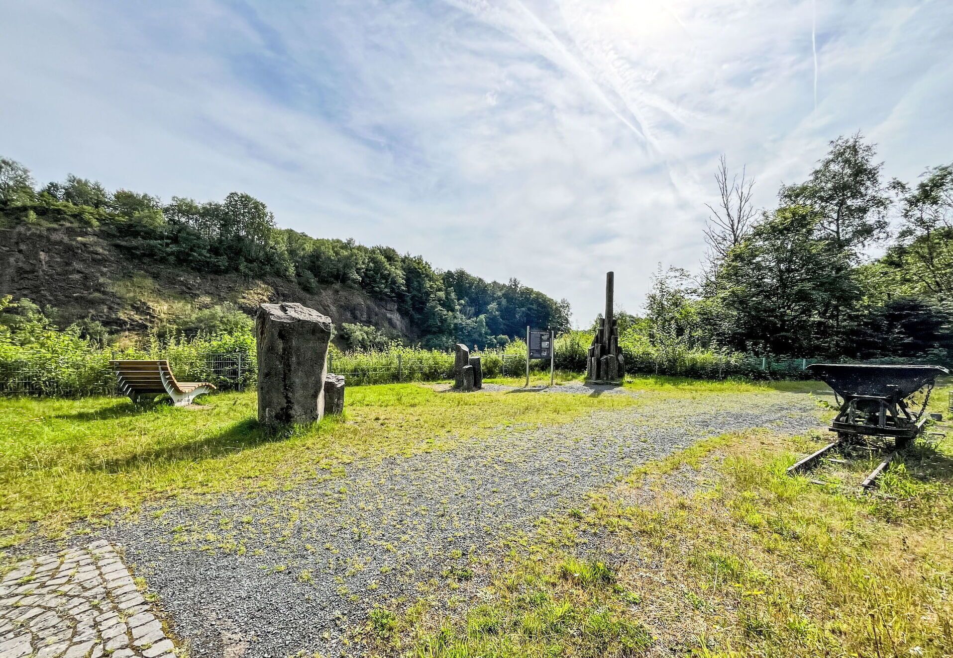 Un sentiero sterrato conduce a una panchina in pietra e una costruzione in pietra, immersi nel verde sotto un cielo sereno.