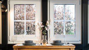 A dining table set with plates, glasses, and a vase of flowers, positioned between two windows with a view of trees.