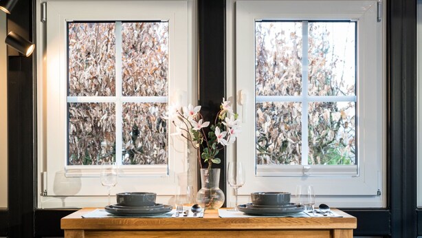 Una mesa de comedor dispuesta con platos, vasos y un florero, ubicada entre dos ventanas con vista a árboles.