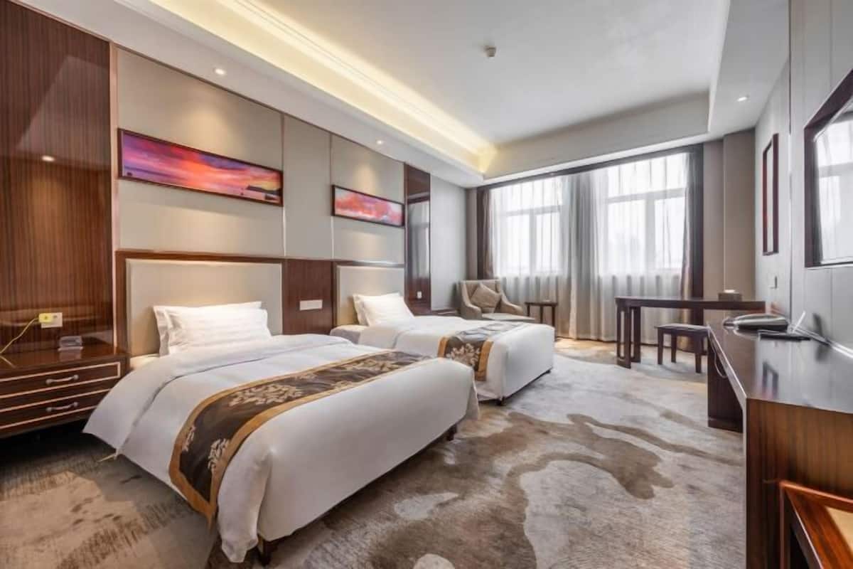 Habitación de hotel con dos camas, un escritorio, una silla, un ventanal grande con cortinas y un televisor montado.