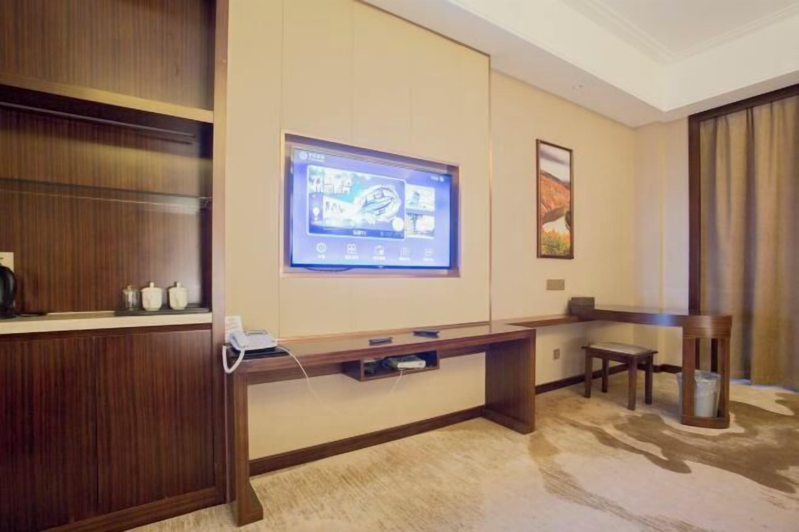 A hotel room with a wooden desk, a flat-screen TV mounted on the wall, a small table, and a trash can.