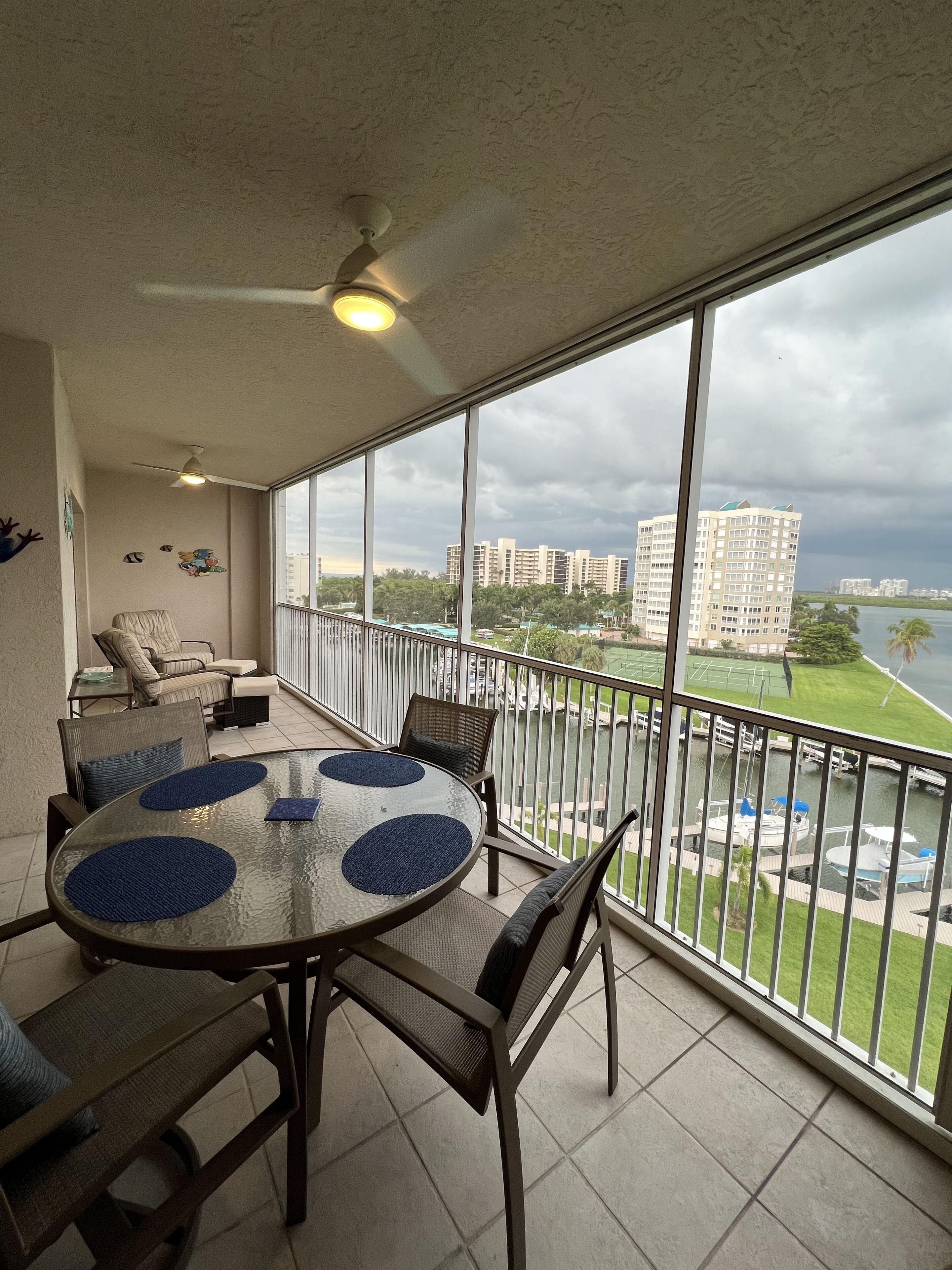A balcony with a glass table and chairs, a ceiling fan, and a view of a marina and buildings.
