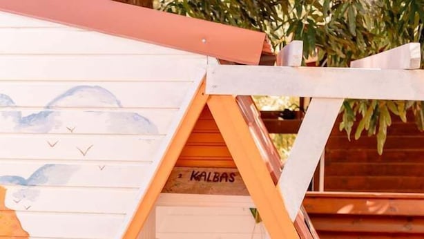 A triangular wooden structure with a bed inside, a door, and a sign that reads "KALBAS". - TIPI 2 Kalbas • Domaine de Meilly (Saint-Louis)