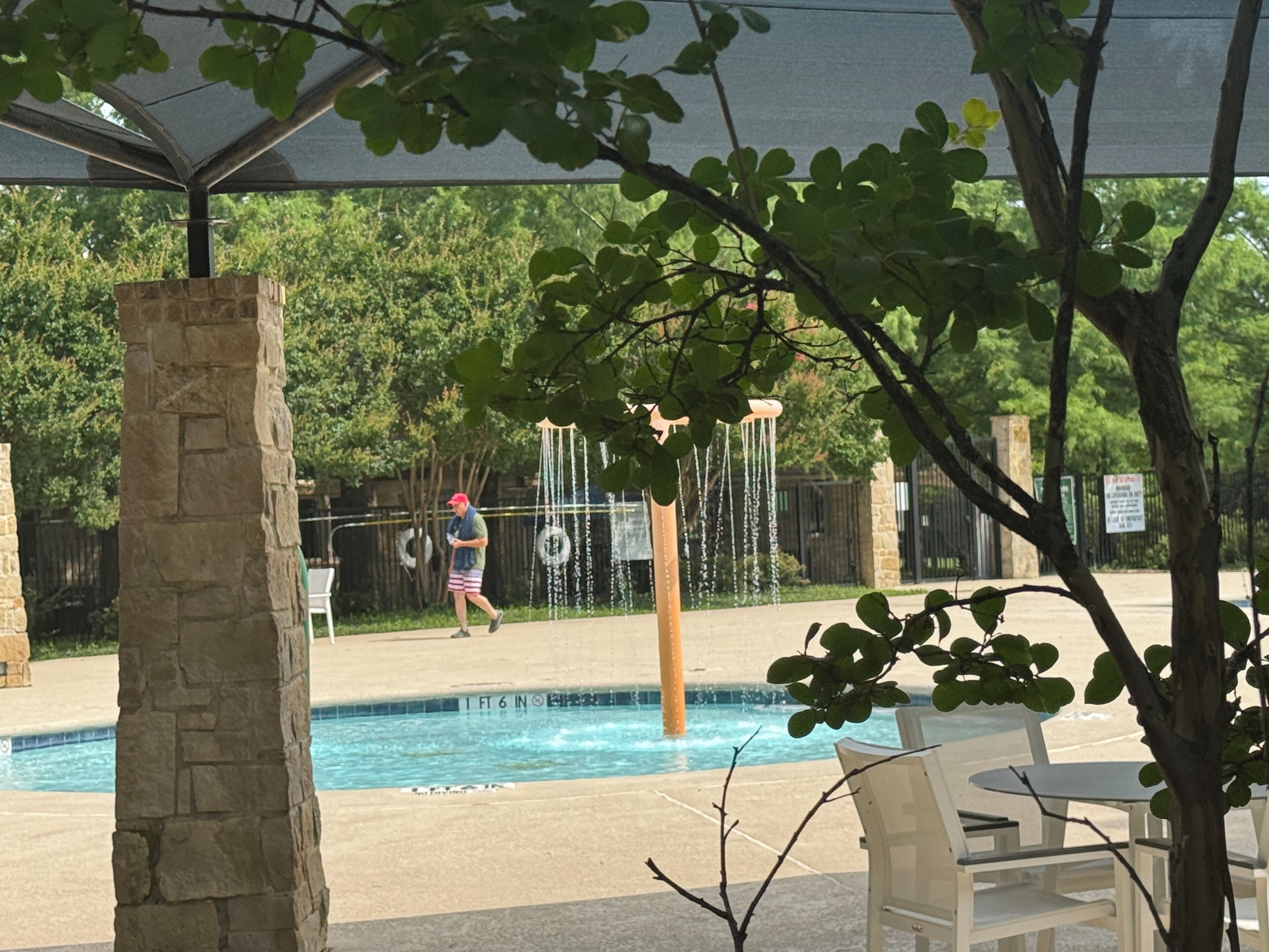 Zona de piscina con un pilar de piedra, un área de descanso cubierta y una persona caminando.