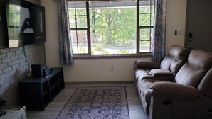 Un salon avec un canapé en cuir, un téléviseur à écran plat fixé à un mur en briques, une grande fenêtre avec des rideaux et un tapis à motifs sur le sol.