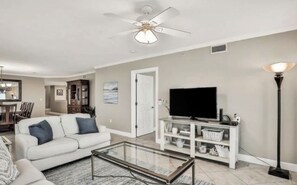 Una sala de estar con un sofá blanco, una mesa de centro de vidrio, un televisor de pantalla plana sobre un soporte blanco, un ventilador de techo y una lámpara de pie.