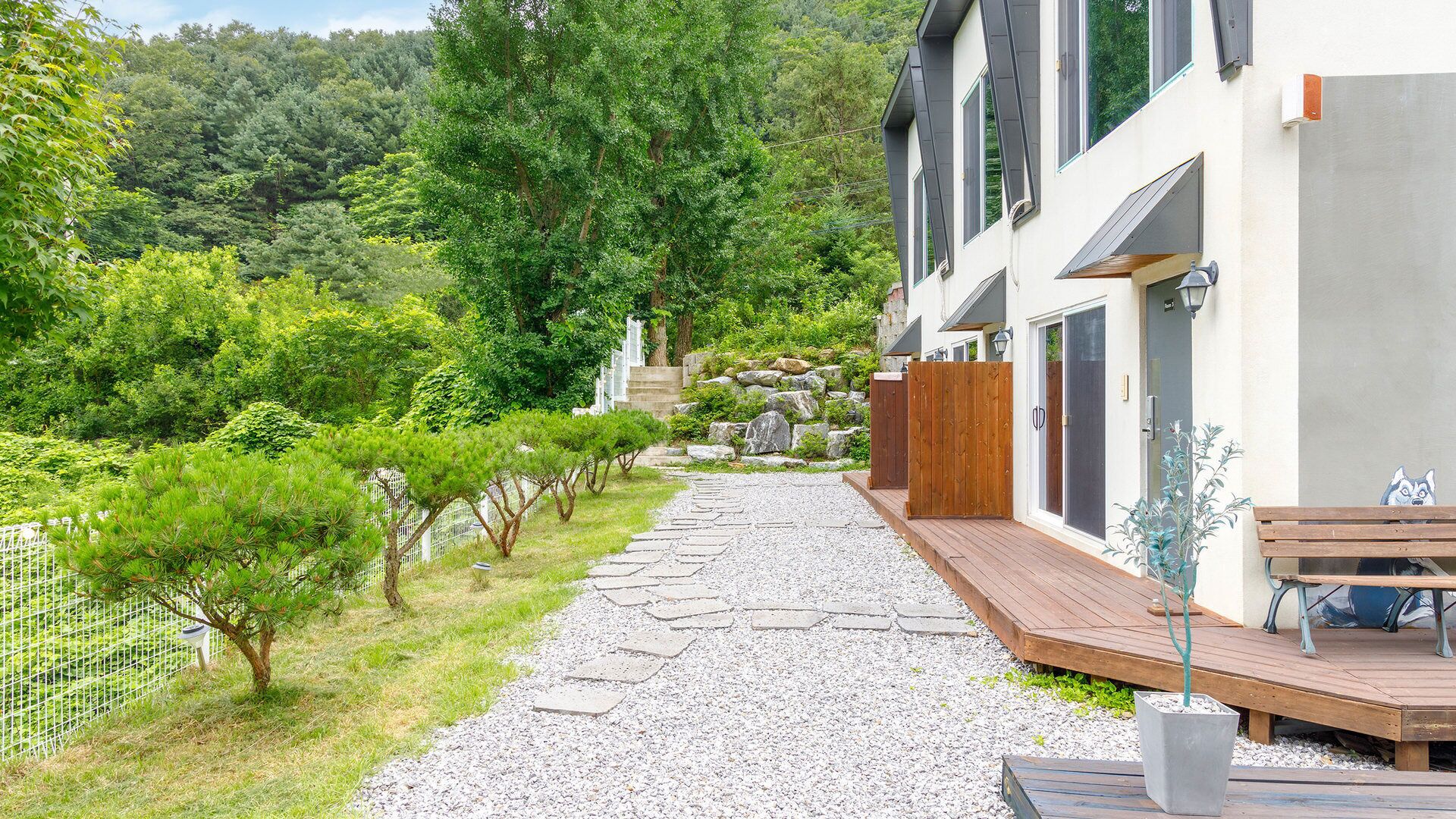 A pathway leading to a modern house with a wooden deck, surrounded by greenery and a stone wall.