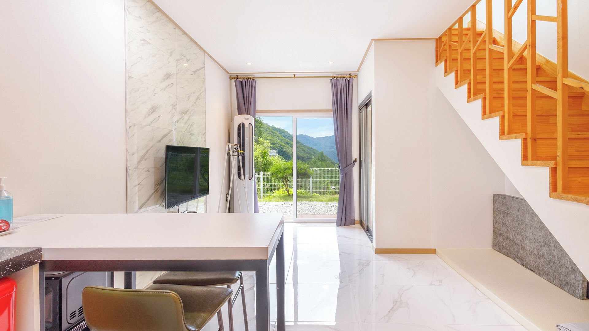 A modern interior with a dining area, a TV, and a staircase leading to a balcony with a mountain view.