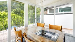 Een eetgedeelte met een tafel met glazen blad, vier houten stoelen en grote ramen met uitzicht op groen.