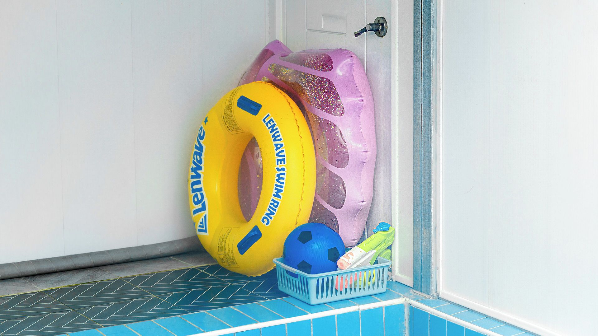 Un espace aménagé au bord de la piscine, avec un sol carrelé de bleu, un flotteur jaune en forme d’anneau, un ballon bleu et un panier en plastique contenant divers objets.