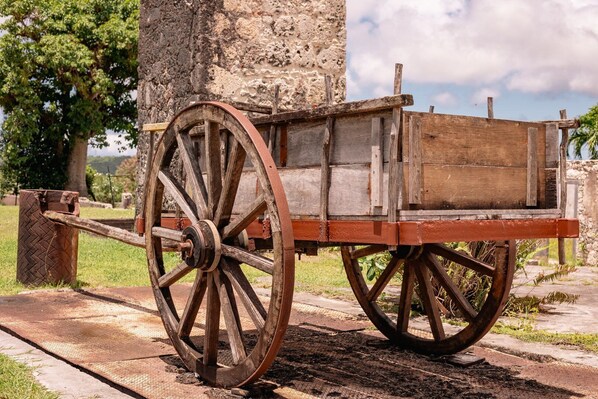 An old wooden cart with large wheels, positioned on a stone path next to a tree and a stone wall.