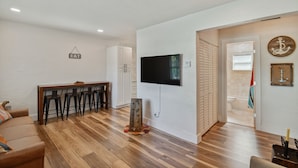A living room with a wooden floor, a sofa, a TV mounted on the wall, and a door leading to a bathroom. - Charming Centrally Located, 3 bedroom house! (Tampa)
