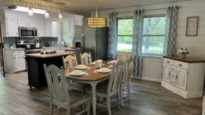 A modern kitchen with white cabinetry, a central island, and a dining area with a wooden table and white chairs. - Cheerful, relaxing ranch in quiet neighborhood with fire pit (Newton)