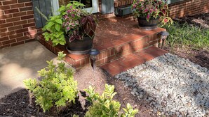 A brick house with a front porch, two potted plants, and a small garden area with mulch and gravel. - Cheerful, relaxing ranch in quiet neighborhood with fire pit (Newton)