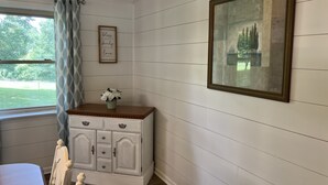 A room with a white paneled wall, a framed picture, a cabinet with drawers, a window with a blue curtain, and a view of greenery outside. - Cheerful, relaxing ranch in quiet neighborhood with fire pit (Newton)