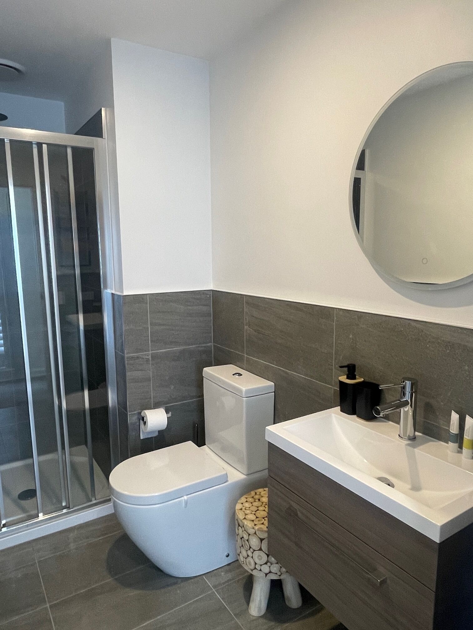 Une salle de bain moderne avec une douche à parois de verre, des toilettes, un lavabo avec un miroir et un petit tabouret en bois.