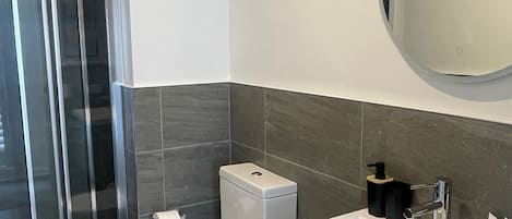 A modern bathroom with a glass shower enclosure, a toilet, a sink with a mirror, and a small wooden stool.