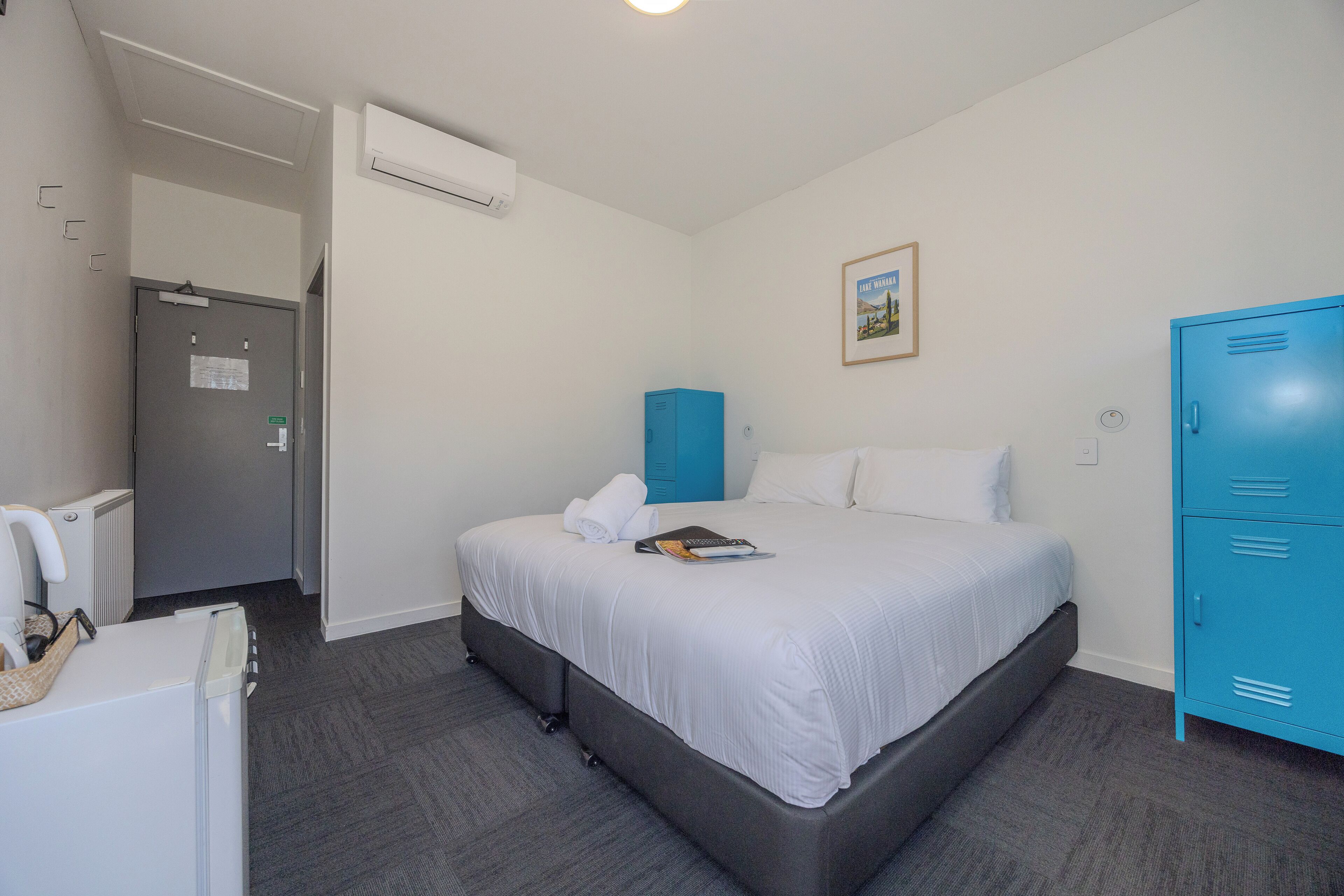 A hotel room with a bed, a blue wardrobe, a white radiator, and a door with a sign.