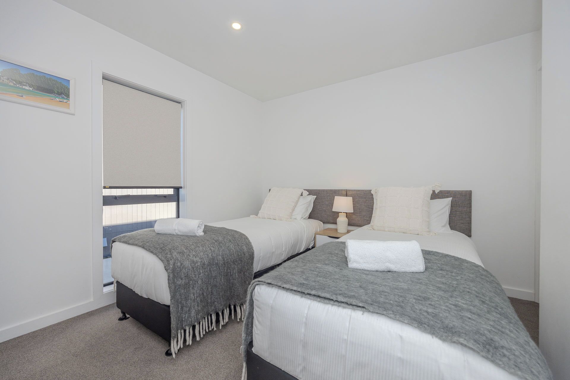 A bedroom with two beds, a window with blinds, a bedside table with a lamp, and a painting on the wall.