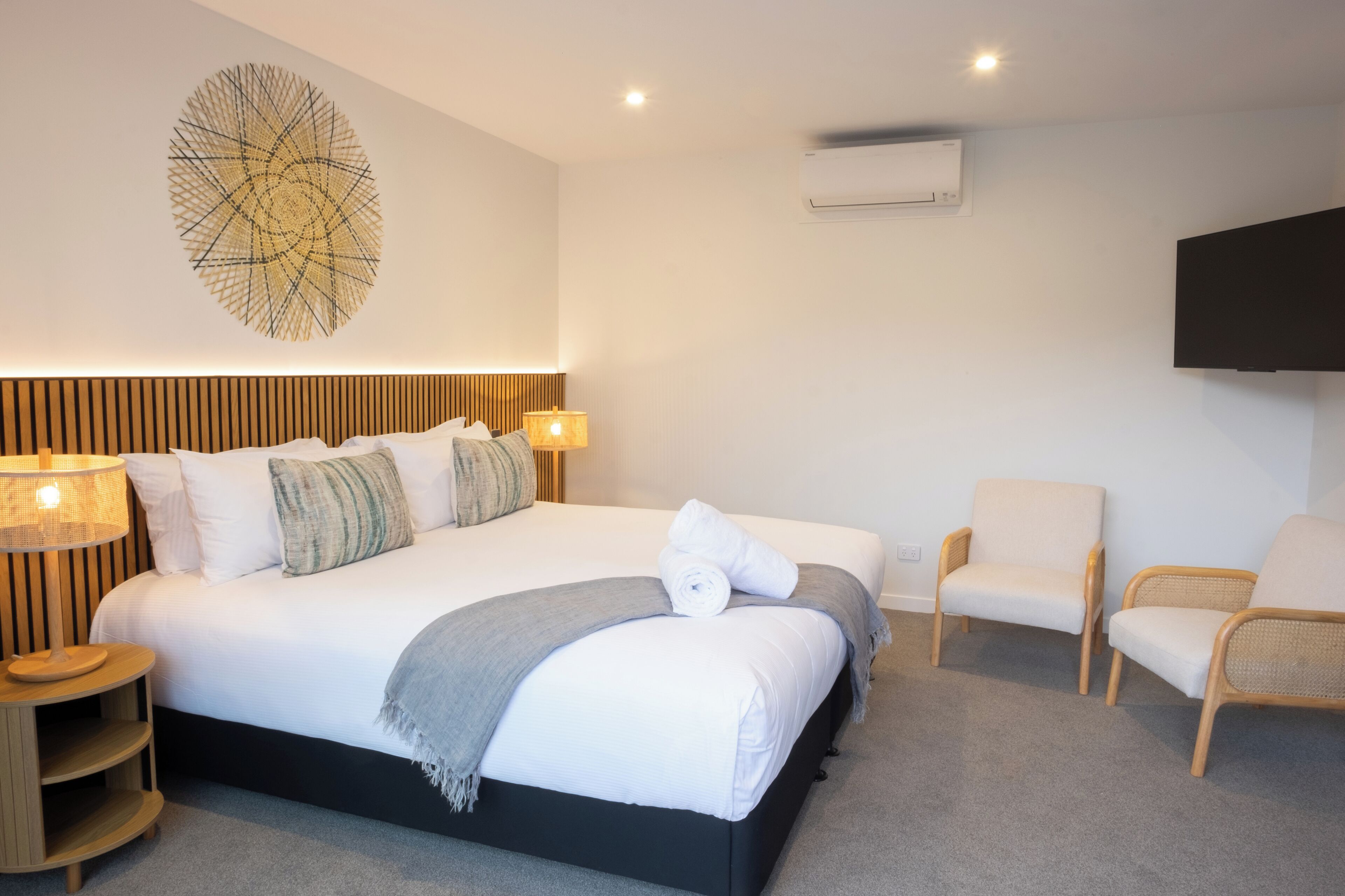 Una habitación de hotel moderna con una cama grande, dos mesitas de noche con lámparas, un televisor montado en la pared y una obra de arte abstracta de gran tamaño.
