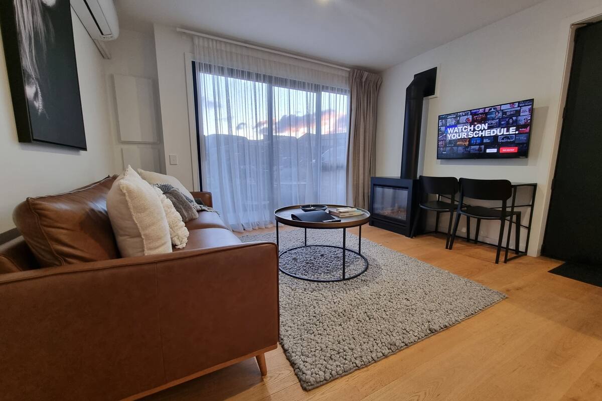 A living room with a brown leather sofa, a small round table, a television mounted on the wall, and a large window with sheer curtains.