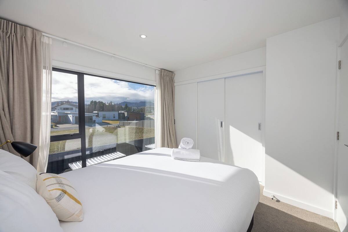 A hotel room with a large bed, a view of buildings and mountains, and a wardrobe.