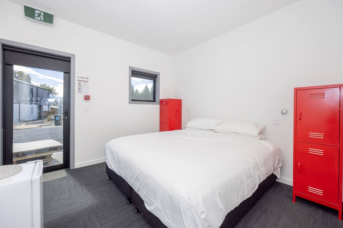 A hotel room with a bed, a red storage unit, a window, and a door leading outside.