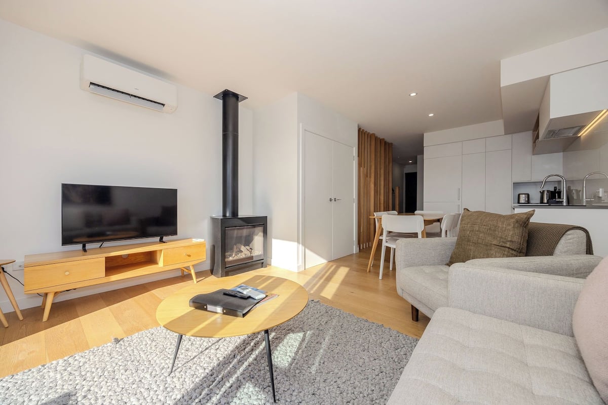 A modern living room with a flat-screen TV, a wooden TV stand, a round coffee table, a sofa, and a wood-burning stove.