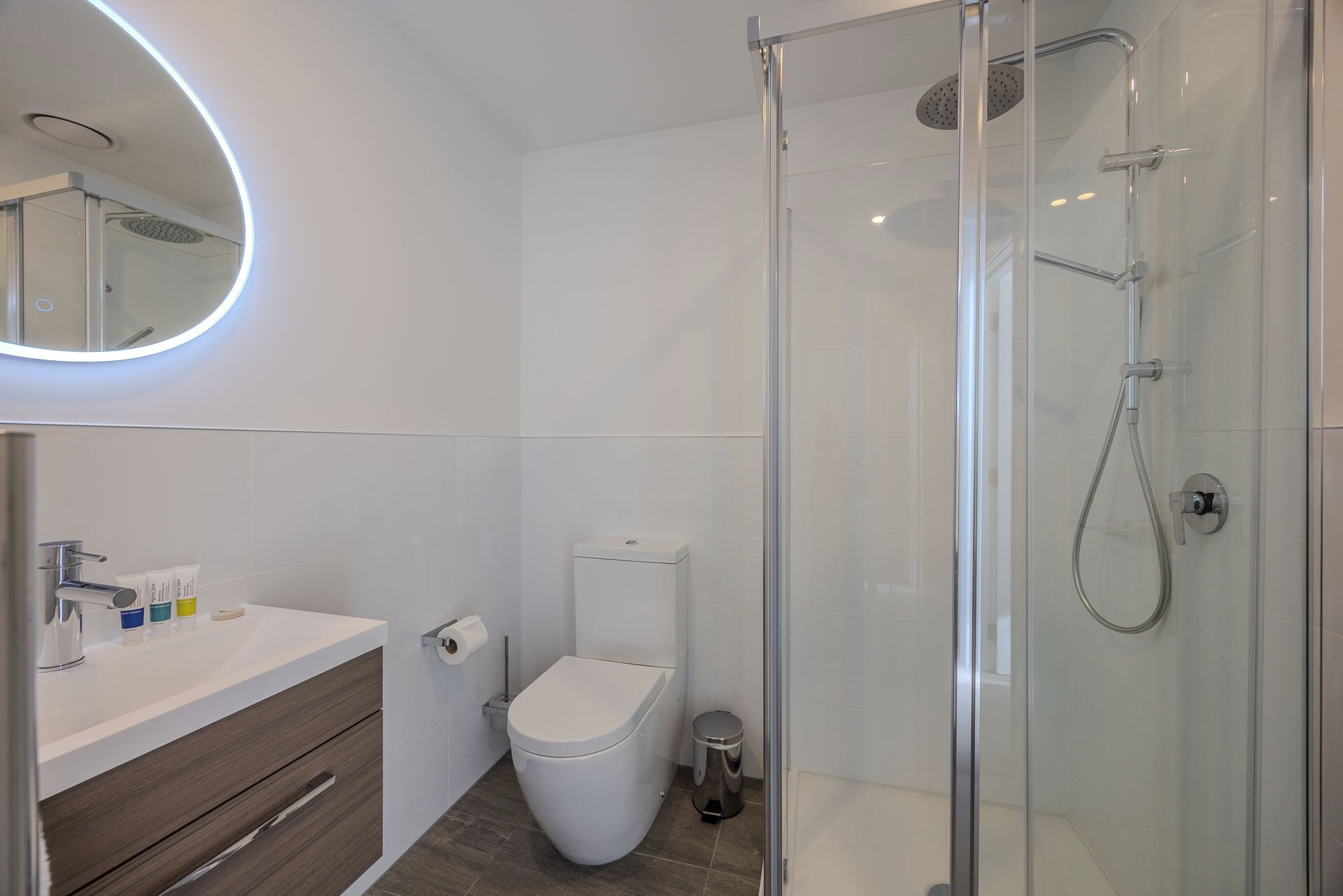 Un baño moderno con ducha de vidrio, inodoro blanco y tocador con espejo y artículos de aseo.