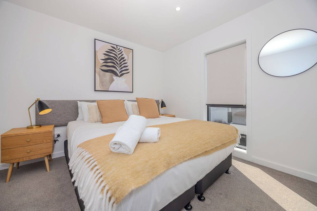 A modern bedroom with a large bed, a bedside table with a lamp, a wall-mounted mirror, and a window with blinds.