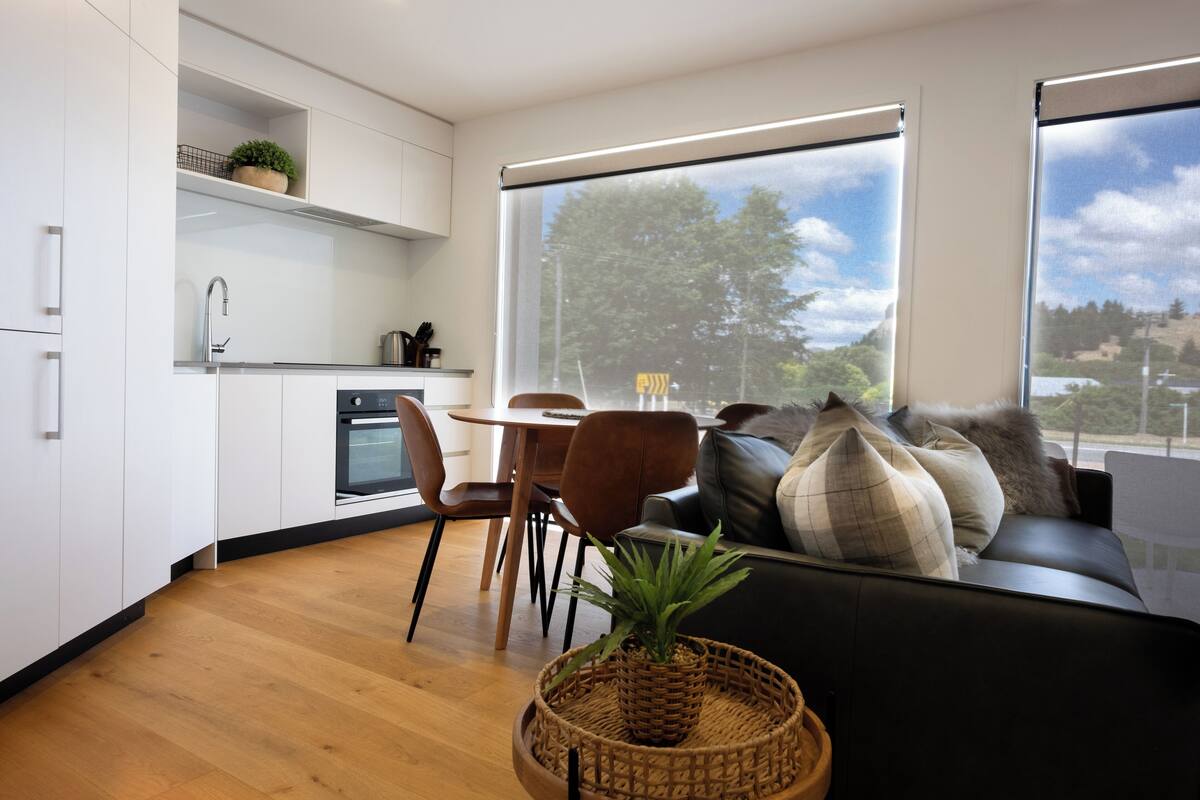 A modern kitchen with white cabinets, a dining area with a wooden table and chairs, and a black sofa with decorative pillows.
