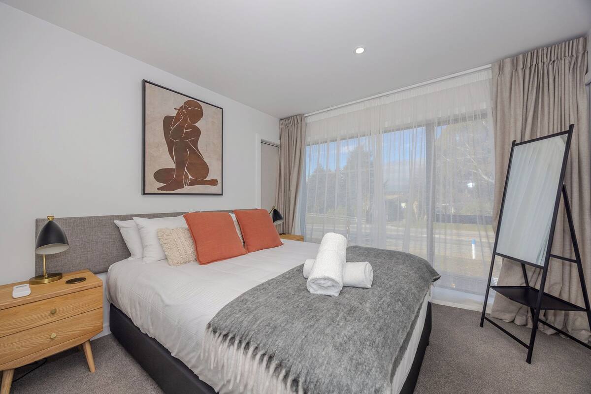 A hotel room with a bed, a bedside table with a lamp, a large window with curtains, and a framed artwork on the wall.