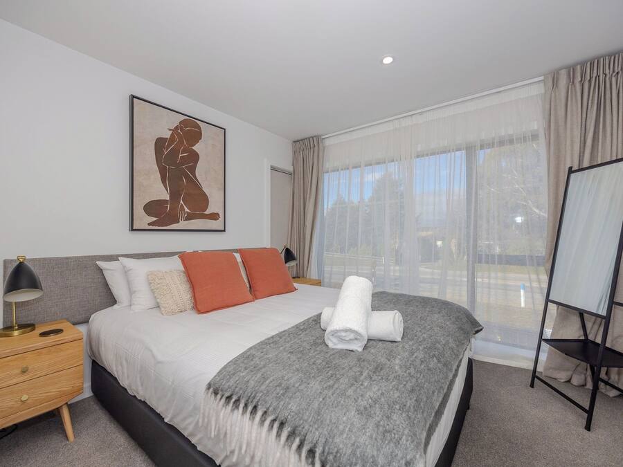 A hotel room with a bed, a bedside table with a lamp, a large window with curtains, and a framed artwork on the wall.
