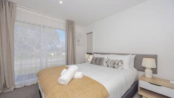 Habitación de hotel ordenada con una cama grande, una mesita de noche con lámpara y vistas a través de cortinas traslúcidas.