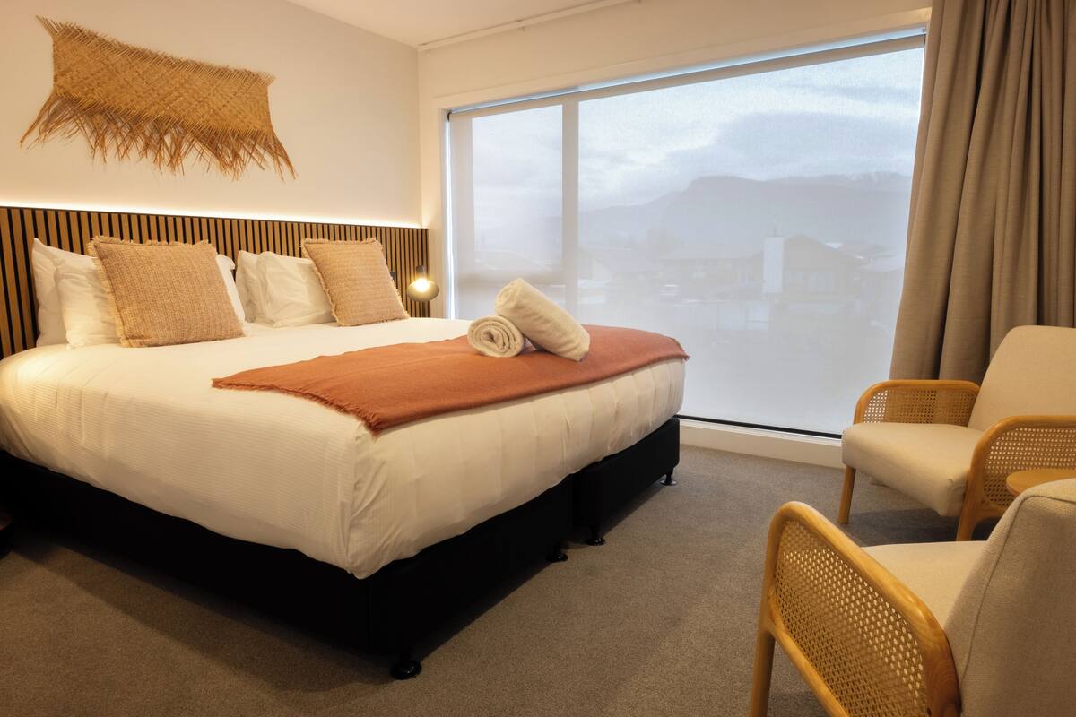 A modern hotel room with a large bed, a view of the city, and a woven wall hanging.